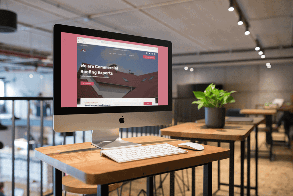 An iMac on a wooden table displaying a "Commercial Roofing Experts" website. The modern office space is softly lit, with a potted plant on a table.