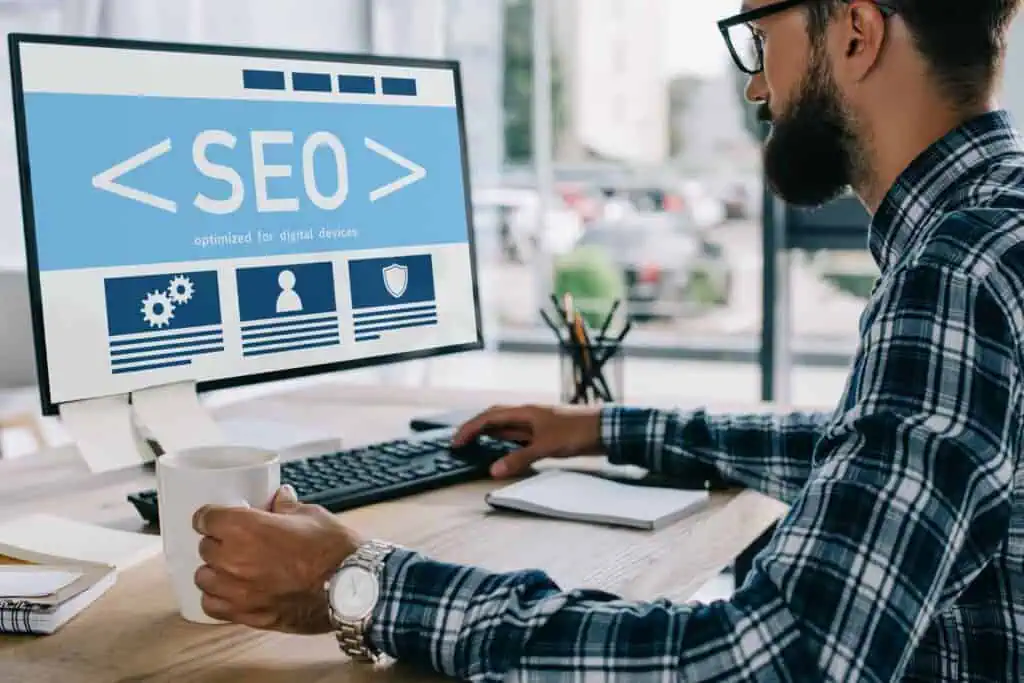 A person with a beard and glasses sits at a desk, facing a computer screen displaying a blue SEO interface. They hold a mug in one hand and have a keyboard and notepad nearby. The workspace is bright and modern.