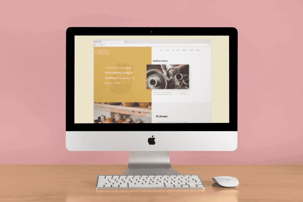 A computer on a wooden desk displays a pottery website. The screen shows handcrafted pots and text about a potters' community. Soft pink wall in the background.