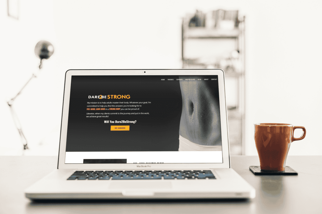 A laptop displaying the "Dare to Be Strong" fitness website with motivational text next to a red mug on a coaster. The setting is minimal and professional.