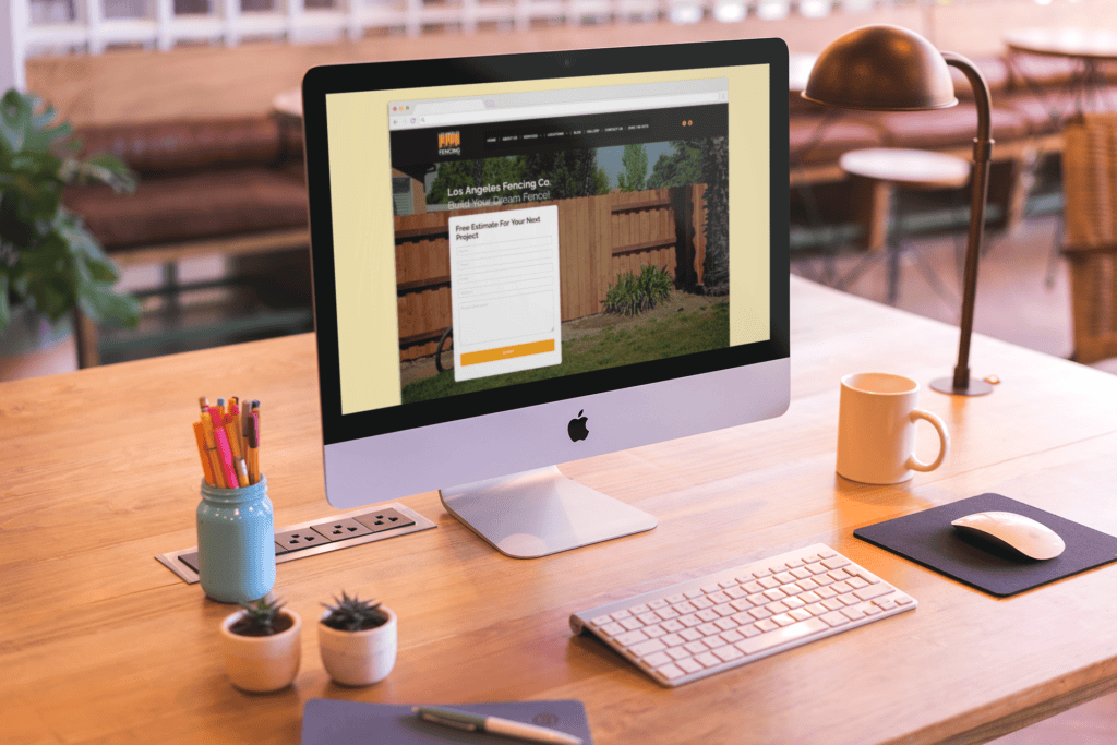 A modern workspace with a sleek iMac displaying a fencing company website. The wooden desk features a keyboard, mouse, lamp, mug, and colorful pencil jar.