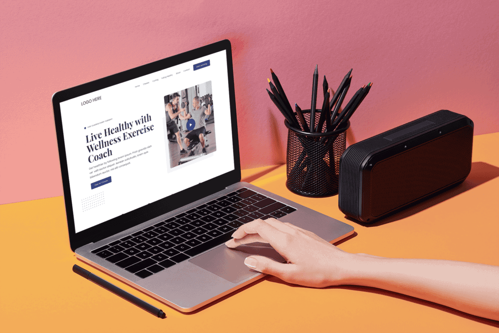 A hand interacts with a laptop on a desk, displaying a wellness coaching website. Nearby are a black speaker and a pencil holder. The setting is colorful and organized.