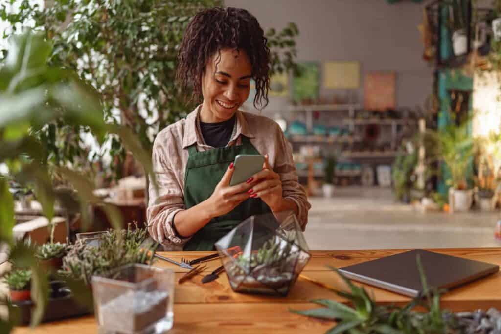 A person with curly hair sits at a wooden table in a plant-filled room, smiling at their phone. They are wearing a green apron and surrounded by small plant arrangements and a laptop.