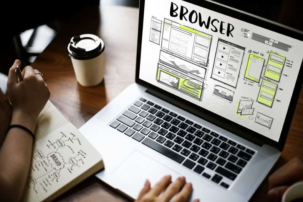 A laptop displays a web design mockup with various layout sketches. Nearby, a person writes in a notebook with a mind map labeled "BRAND." A coffee cup sits by their side.
