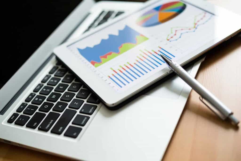 A tablet displaying colorful graphs and charts rests on a laptop keyboard. A pen is placed beside the tablet. The images on the tablet show bar charts, pie charts, and line graphs, suggesting data analysis or a business presentation.